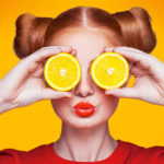 Women with red hair, lips and shirt holding orange slices over eyes on bright orange background