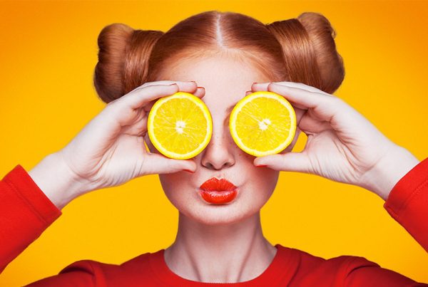 Women with red hair, lips and shirt holding orange slices over eyes on bright orange background