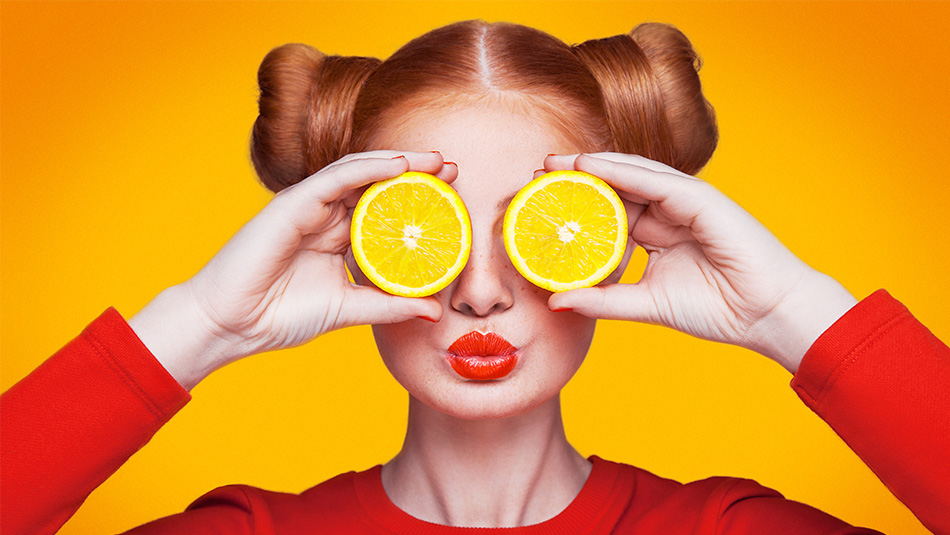 Women with red hair, lips and shirt holding orange slices over eyes on bright orange background