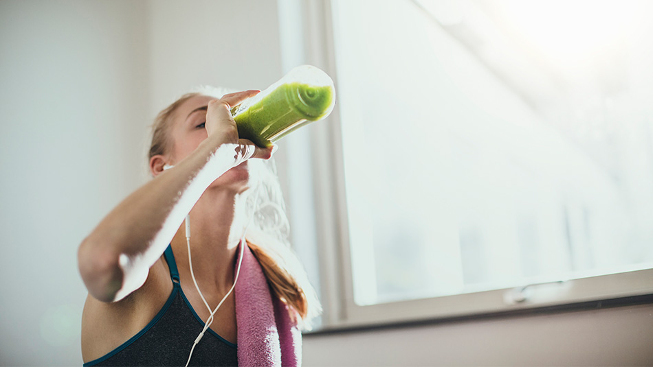 Girl in a workout studio with pink towel on her shoulder drinking green drink out of water bottle listening to music with ear buds