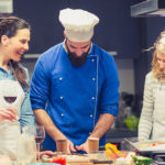 Chef in blue coat instructing two women students on prepping food