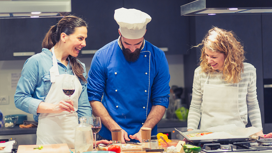 Chef in blue coat instructing two women students on prepping food