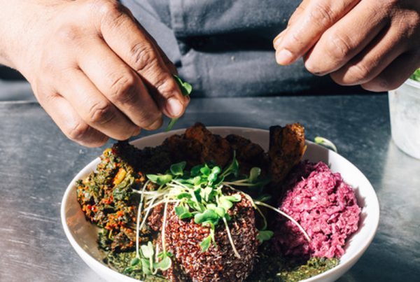 White plate of colorful food on metal table with hands garnishing