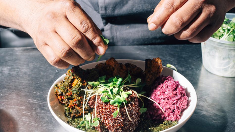 White plate of colorful food on metal table with hands garnishing